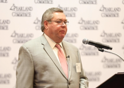 A mid shot to a man in a conference. The man appears to be 50 years old wearing a grey suit and is speaking standing in front of a microphone.