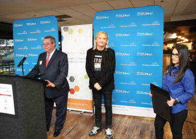 A picture of the guitarist Joe Walsh stanting in a conference. Beside him, there is a man in suit talking to the microphone and in the other side there is a woman in a formal blouse carrying what appears to be a commemorative plaque.