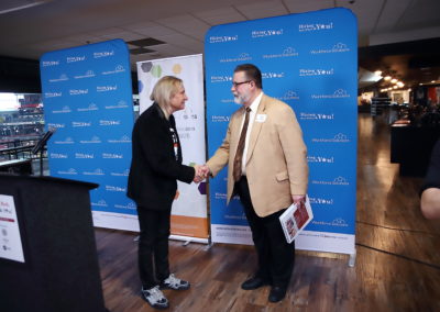 A picture of the guitarist Joe Walsh and a man shaking hands. Walsh is wearing a casual suit and sneakers and the other is wearing a formal light brown suit and has short hair and beard and is holding a book in his free hand. The people are shaking hands in front of some ads with the “Workforce Solutions” logo and a “Hiring Red, White & You” logo.