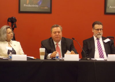 A close up to members of a corporative roundtable meeting. There is two man and a woman in formal clothes. They have coffee, water, and microphones, they seem to be engaged in a discussion.