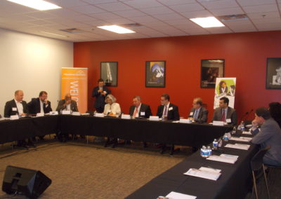 A group of people sitting at a round table conference engaged in a discussion. They appear to be diverse in age, gender, and ethnicity. The table is covered with papers, notebooks, and pens. The room has posters in the back and appears to be a conference room.